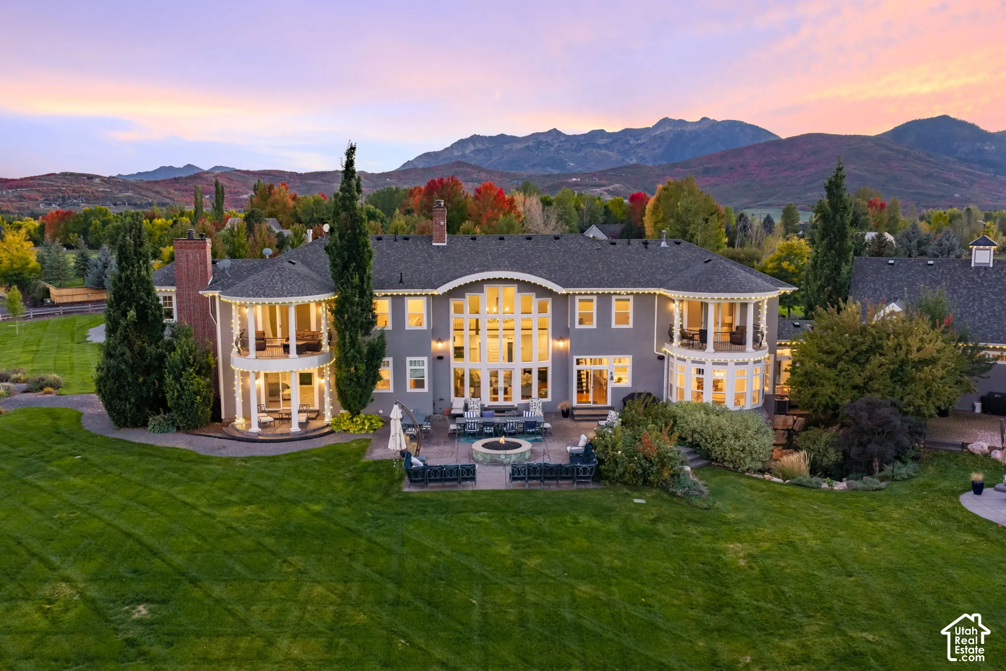 Multi-story residence featuring dual-level semi-circular balconies, expansive floor-to-ceiling windows, stone-surround patio, built-in circular fire feature, and mountain backdrop