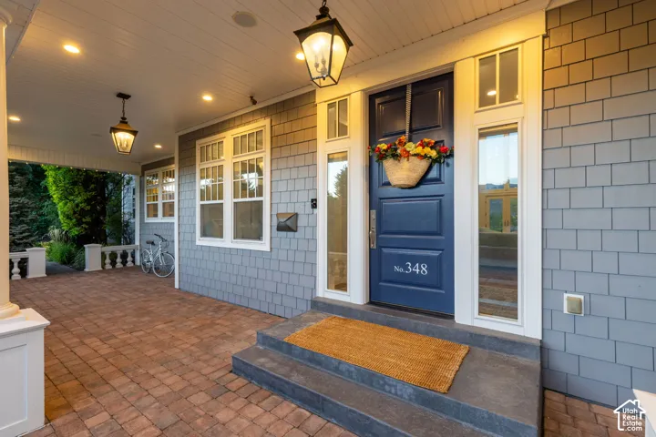 Covered porch featuring brick paver flooring, shingle-style siding, custom blue front door with sidelights, and pendant lighting