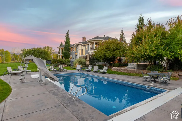 In-ground swimming pool with an integrated slide, expansive concrete patio, tiered stone retaining walls, and mature landscaping