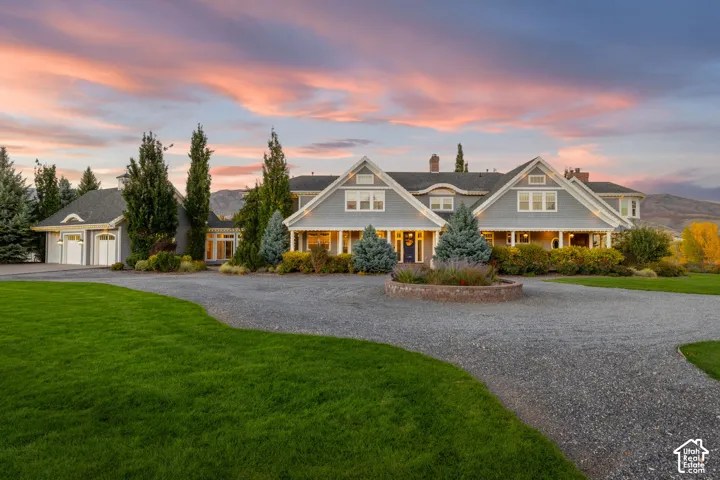 Cape Code-style exterior featuring gray horizontal siding, a covered front porch, a multi-gabled roofline, an expansive gravel motor court, and a circular stone landscape planter