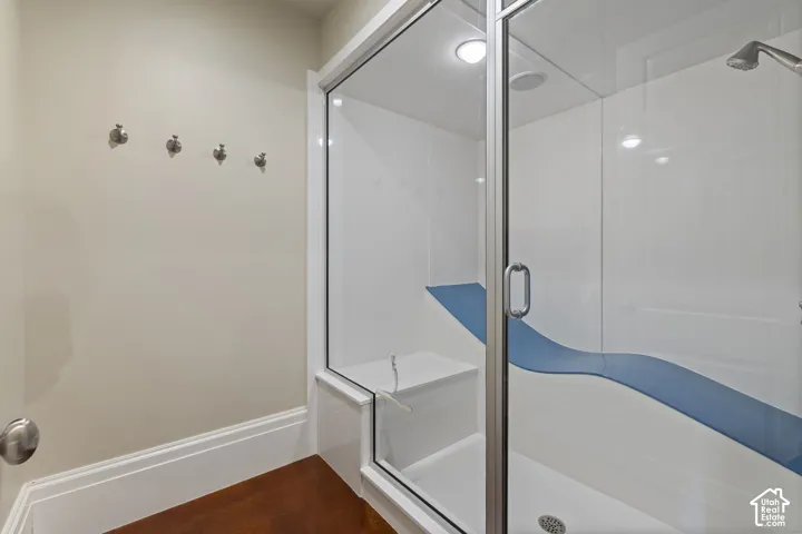 Steam shower enclosure featuring glass doors, white surround panels, an integrated bench seat, and a wall-mounted shower head, finished with wood-finish flooring and four brushed metal wall-mounted hooks