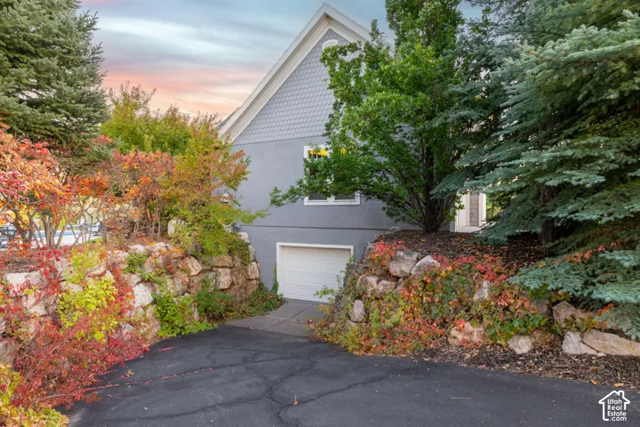 Lower garage entrance featuring a circular asphalt driveway lined with mature landscaping, including vibrant seasonal foliage and established evergreen trees