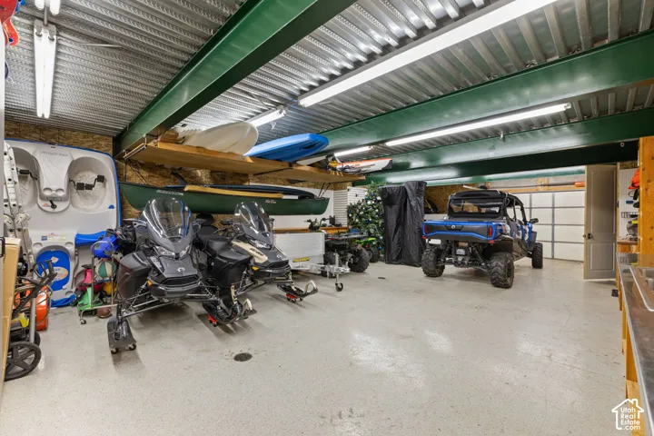 Spacious lower garage featuring high ceilings with corrugated metal finish, painted steel support beams, epoxy-coated concrete flooring, fluorescent strip lighting, and custom built-in wooden storage shelving