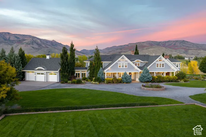 Lakefront estate featuring a multi-gabled roofline, three-car garage, circular gravel driveway, manicured lawn, and expansive mountain backdrop