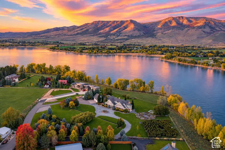 Aerial perspective of a sprawling waterfront estate featuring expansive lawns, mature trees, paved circular driveway, and dramatic mountain backdrop. Accurate AI generated depiction of the reservoir at full capacity. Currently drained for pipeline repair.