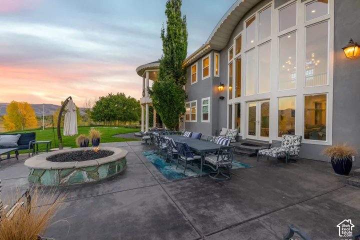 Expansive rear patio featuring concrete hardscaping, a stone-accented fire pit, a curved multi-story exterior with floor-to-ceiling windows, and a covered balcony