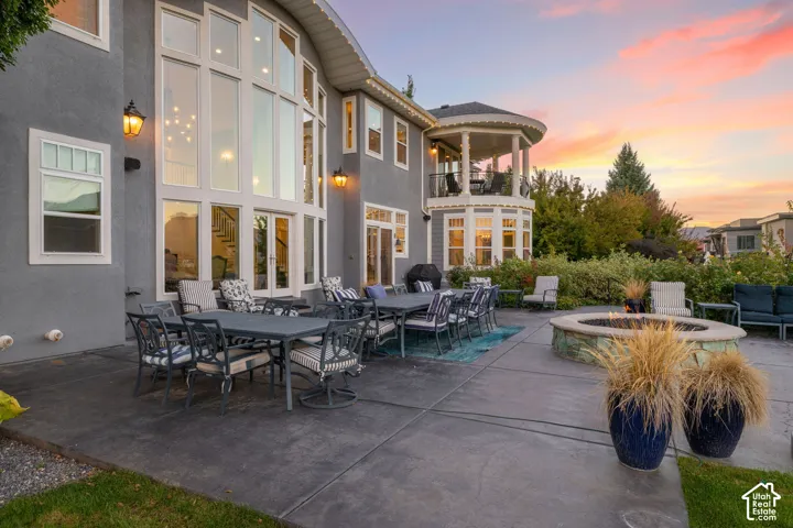 Expansive concrete patio featuring a stone-clad circular fire pit, multi-story floor-to-ceiling windows, second-story covered balcony, and exterior wall-mounted lighting
