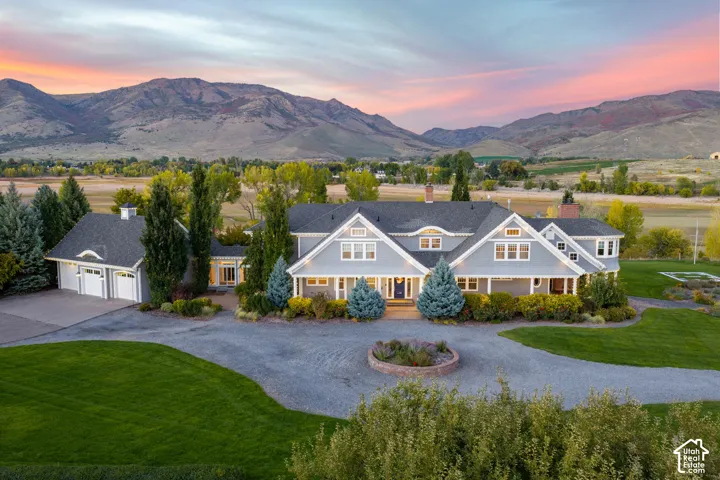 Lakefront residence with grey siding and stone exterior accents, featuring a detached multi-car garage, circular driveway, manicured landscaping, and mountainous backdrop