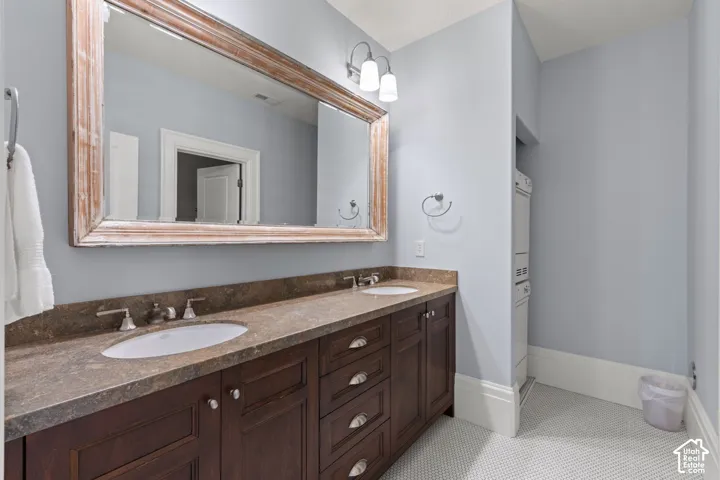 Dual-sink vanity with dark wood cabinetry, stone-finish countertops, wall-mounted light fixture, and large framed mirror, stacked washer and dryer