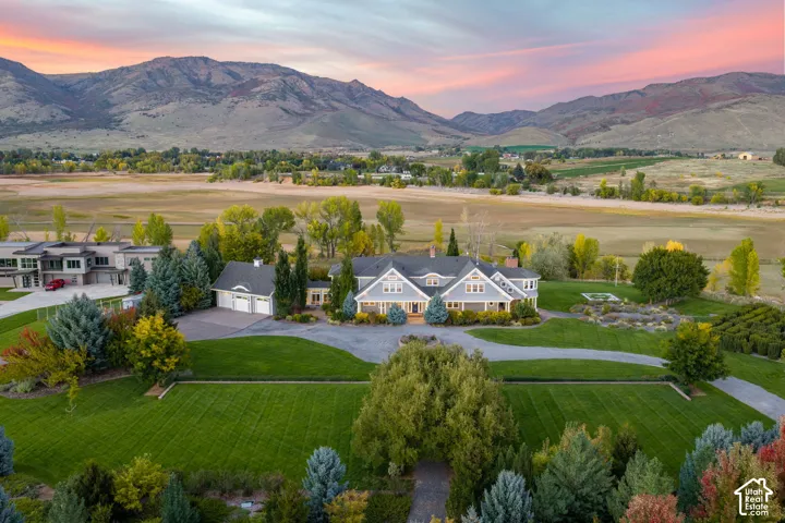 Aerial perspective of a lakefront estate featuring a shingled roof, manicured lawn, paved circular driveway, detached garage, and mountain backdrop