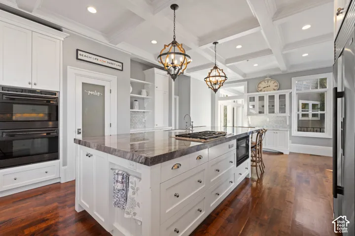 Gourmet kitchen featuring a coffered ceiling, center island with stone countertop and gas cooktop, double wall ovens, wood-finish flooring, and pendant lighting