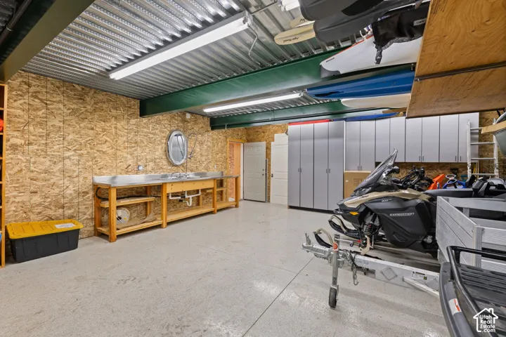Lower garage workspace featuring a stainless steel countertop, integrated sink, built-in floor-to-ceiling storage cabinetry, exposed corrugated metal ceiling, and linear fluorescent lighting