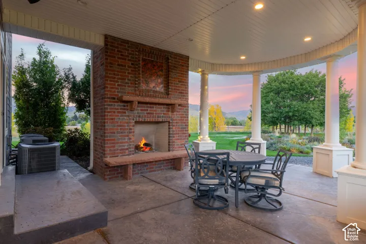 Covered patio featuring a brick fireplace with a stone hearth, recessed lighting, and white structural columns overlooking an expansive landscape