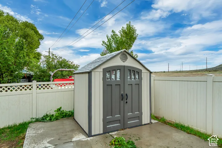 Storage shed with double doors, paneled siding, shingled roof, and integrated windows, situated on a concrete pad and enclosed by perimeter vinyl fencing