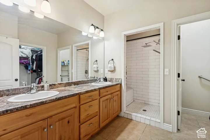 Bathroom featuring a dual-sink vanity with stone-finish countertops, full-width mirror, walk-in shower with subway tile surround, and tile flooring
