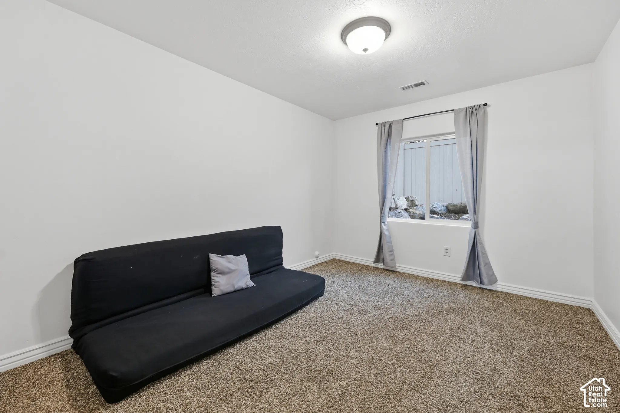 Neutral-toned room featuring neutral wall-to-wall carpeting, white walls, a flush-mount ceiling light fixture, and a window with exterior stone landscaping visible