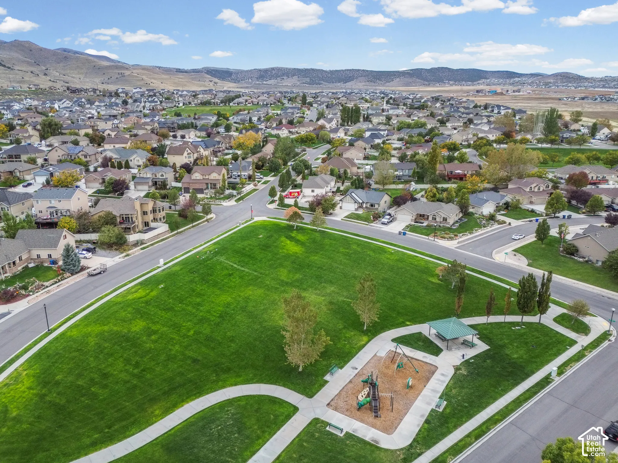 Expansive common green space featuring a manicured lawn, paved walking paths, a shaded pavilion, and a playground, all set against a mountain backdrop