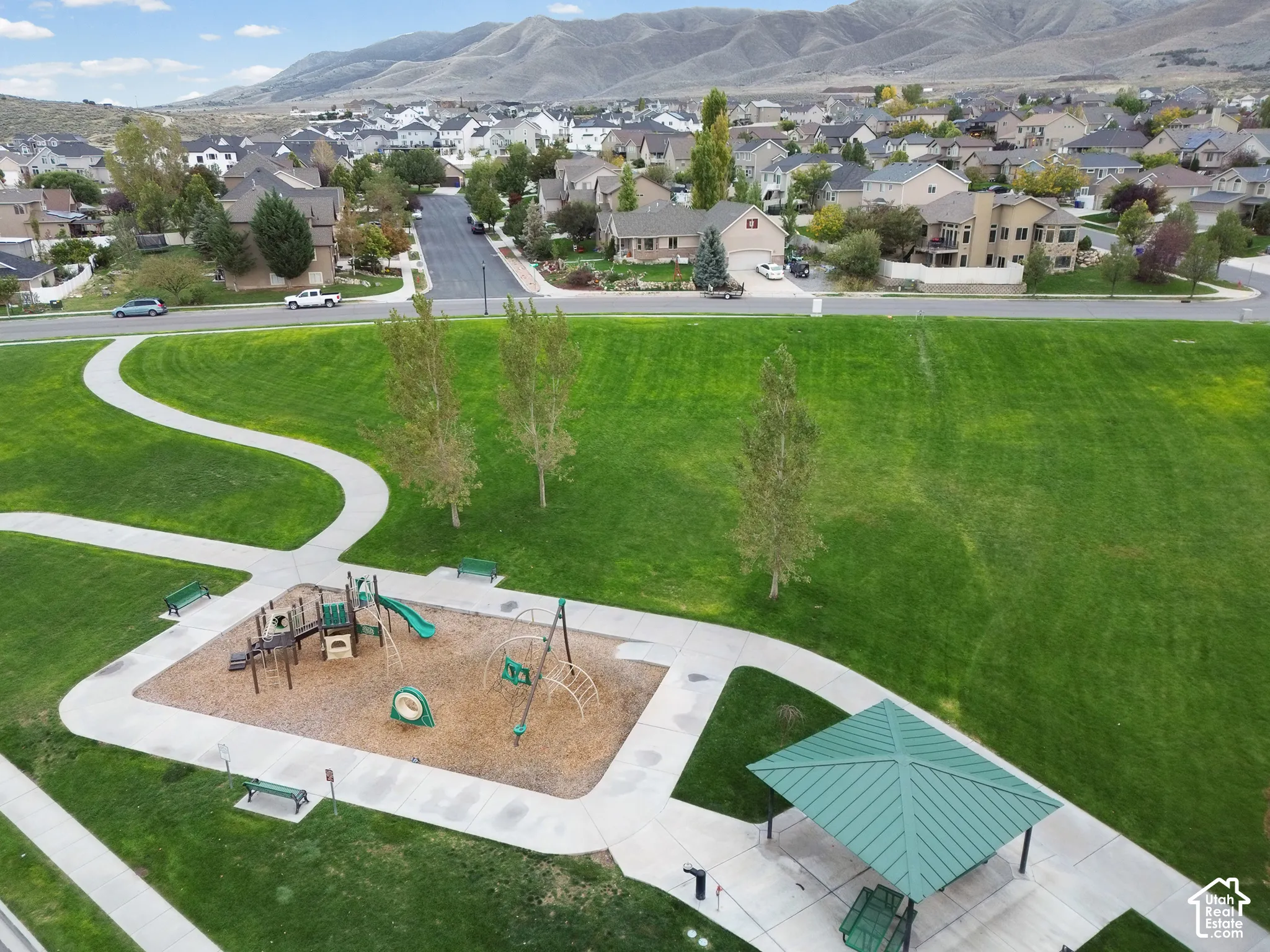 Aerial perspective of a park featuring a manicured lawn, concrete walking paths, a playground structure with engineered wood fiber surfacing, a metal-roofed pavilion, and a mountain backdrop