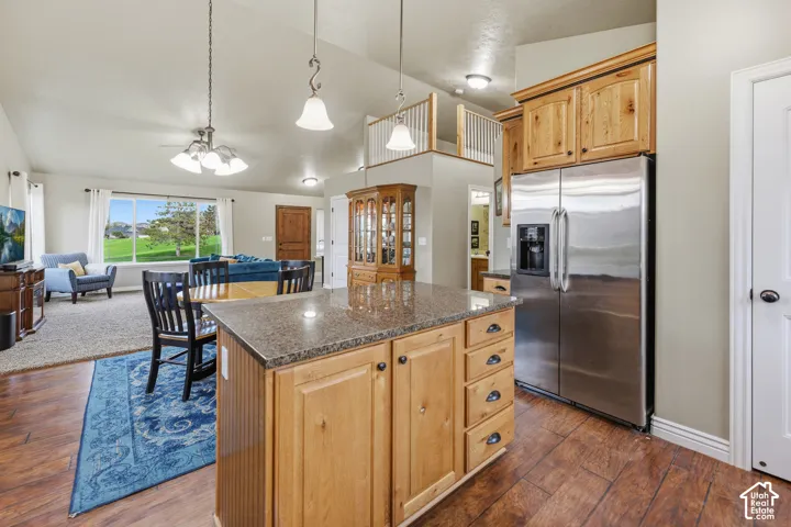 Kitchen featuring a stone-topped center island with storage cabinetry, stainless steel side-by-side refrigerator, wood-finish flooring, and vaulted ceilings with pendant lighting