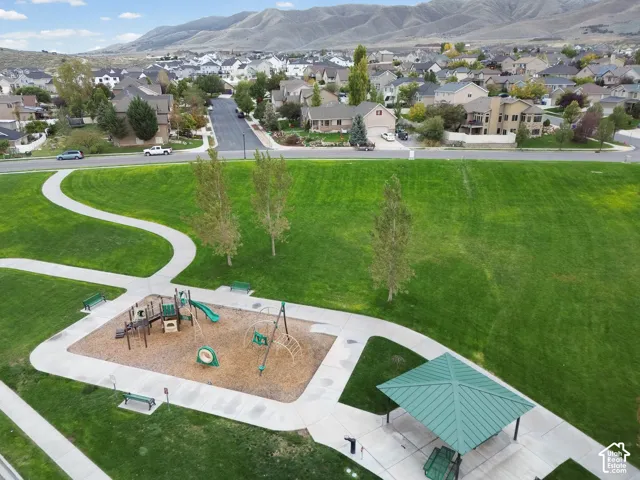 Aerial perspective of a park featuring a manicured lawn, concrete walking paths, a playground structure with engineered wood fiber surfacing, a metal-roofed pavilion, and a mountain backdrop