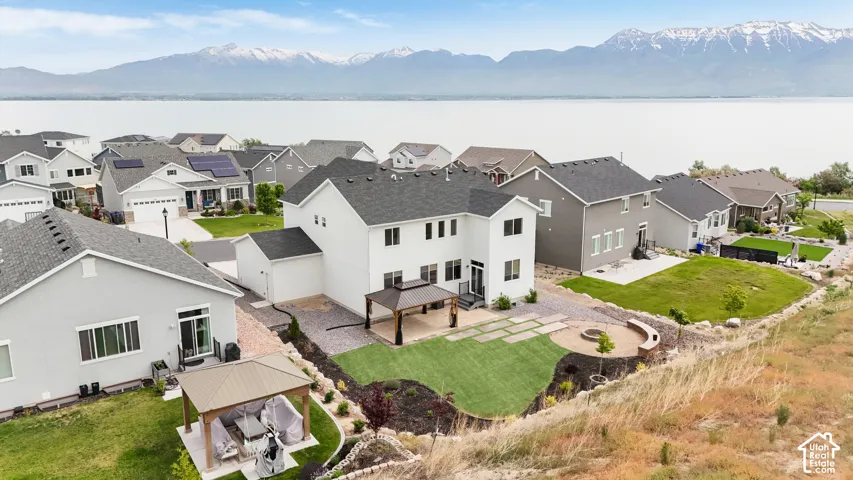 Aerial perspective of a two-story residence featuring a landscaped backyard, a permanent pergola, a circular hardscaped fire pit area with integrated seating, and a backdrop of mountain and water scenery