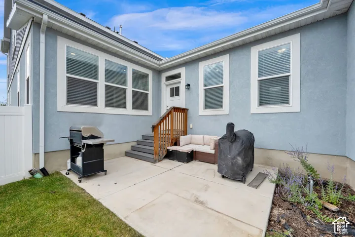 Rear exterior featuring light blue stucco siding, a concrete patio, wood steps with a handrail, multiple pane windows, and white vinyl perimeter fencing