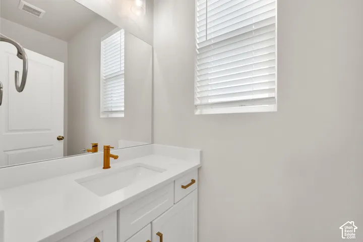 White cabinetry with gold-toned hardware, rectangular undermount sink, solid-surface countertop, full-width mirror, and window with shutter blinds