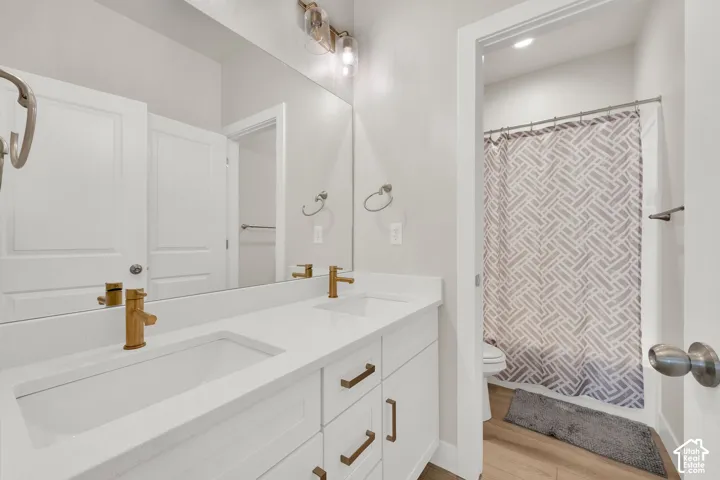 Dual-sink vanity with white cabinetry, light-toned stone countertop, brass-finish fixtures, and full-width vanity mirror