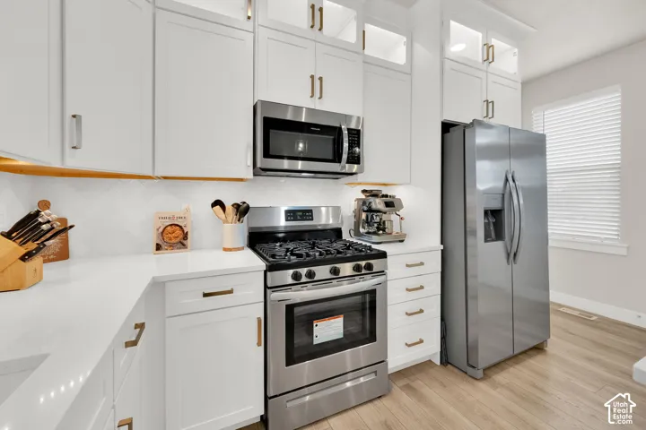 White shaker-style cabinetry with gold-tone hardware, quartz countertops, herringbone tile backsplash, stainless steel appliances, and wood-finish flooring