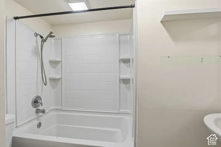 White bathtub with integrated surround, chrome handheld shower fixture, and a built-in wall shelf