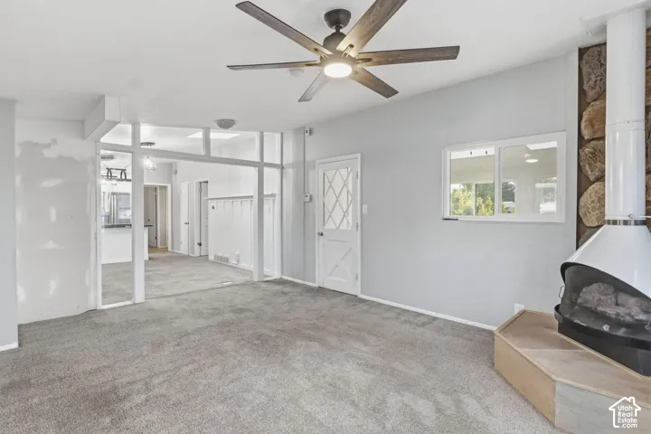 Interior space featuring a white, modern freestanding fireplace with a stone-surround accent wall, wood-finish ceiling fan, wall-to-wall carpeting, and a glass-paneled exterior door