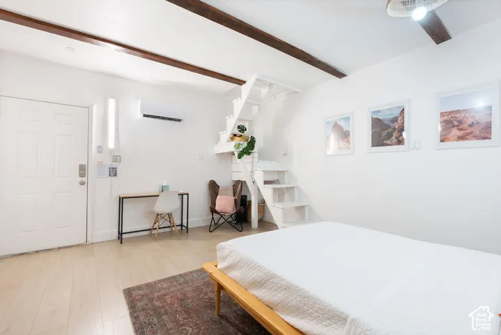 Exposed dark wood ceiling beams, light wood-finish flooring, white staircase with open risers, wall-mounted climate control unit, and a six-panel entry door