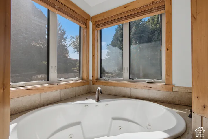 Jetted soaking tub featuring tile surround, wood-trimmed windows, and matte metal fixtures