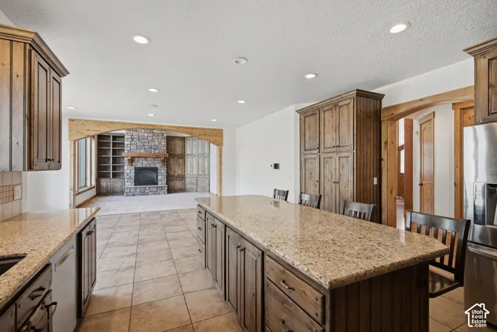 Kitchen featuring stained wood cabinetry, stone countertops, large center island, and tile flooring, with a stone-faced fireplace and built-in shelving visible in the background