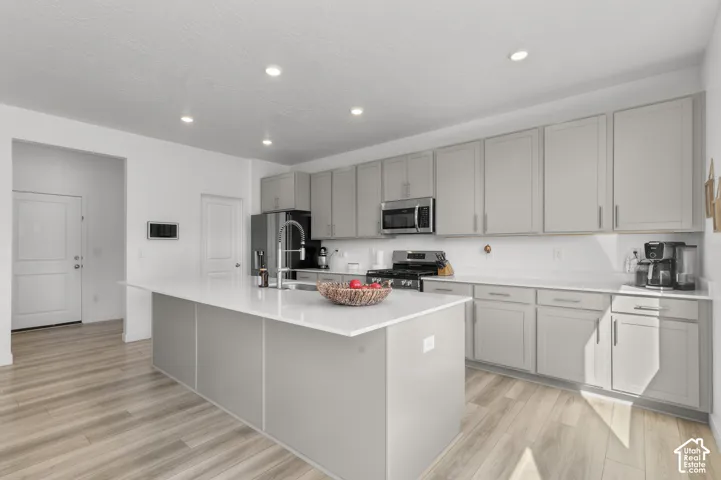 Kitchen featuring light gray cabinetry, expansive quartz countertops, an oversized center island with an integrated sink, stainless steel appliances, and wood-finish flooring