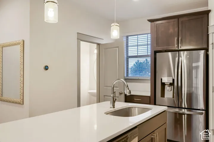 Kitchen featuring an island with a light-toned countertop, under-mount stainless steel sink, stainless steel refrigerator, dark-finish cabinetry, and glass pendant lighting