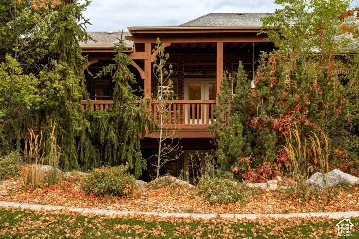 Brick exterior featuring a covered wooden deck with railings, mature coniferous landscaping, and a concrete garden border