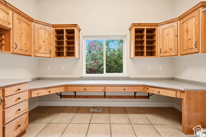 Built-in natural wood cabinetry with integrated shelving and desk-height workspace, topped with neutral solid-surface counters, featuring ceramic tile flooring and a central window