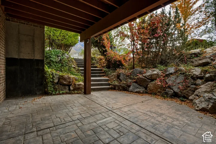 Covered patio featuring paver flooring, timber support beam, tiered stone retaining walls, and concrete stairs