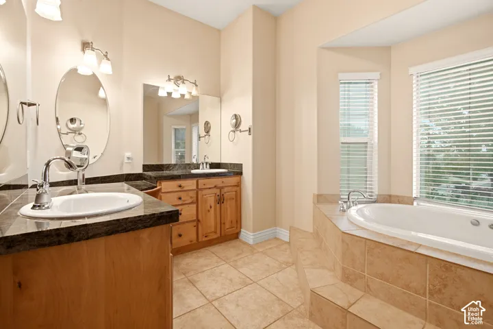 Dual-sink vanity with dark stone countertops, natural wood cabinetry, oversized jetted soaking tub, and ceramic tile flooring