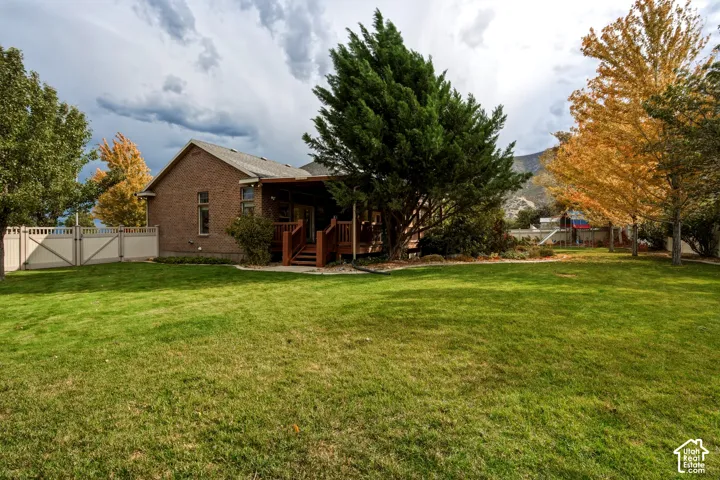 Brick exterior residence featuring a covered wooden deck, expansive lawn, mature trees, and perimeter vinyl fencing