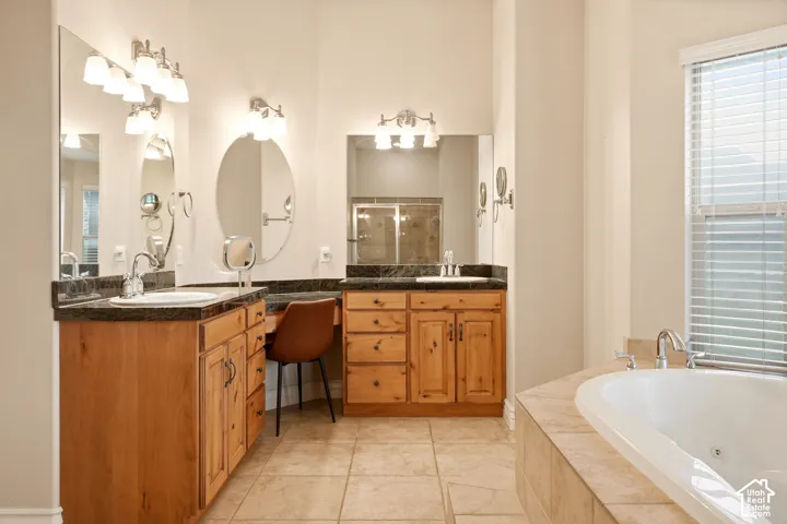 Dual-vanity cabinetry with integrated makeup desk, stone-finish countertops, drop-in sinks, ceramic tile flooring, and a jetted soaking tub with a tiled deck