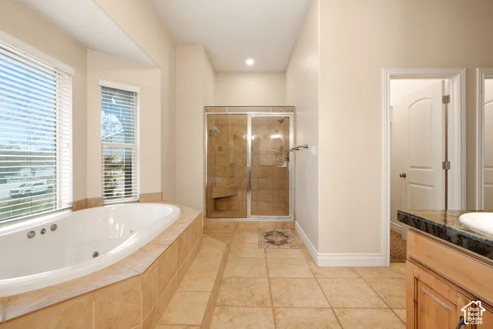 Jetted soaking tub with tile surround, glass-enclosed shower with built-in seating, natural stone-finish floor tiling, dual-paned windows, and vanity with stone countertop