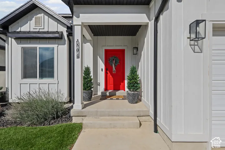 Exterior entry featuring vertical board-and-batten siding, a covered porch, solid-core red front door, and geometric wall-mounted outdoor lighting