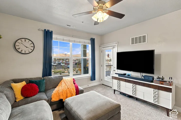 Bright living room showcasing expansive windows with scenic vistas, ceiling fan with integrated lighting, neutral carpeting, and a wall-mounted flat-screen television above a white media console with wood-finish top and storage cabinetry