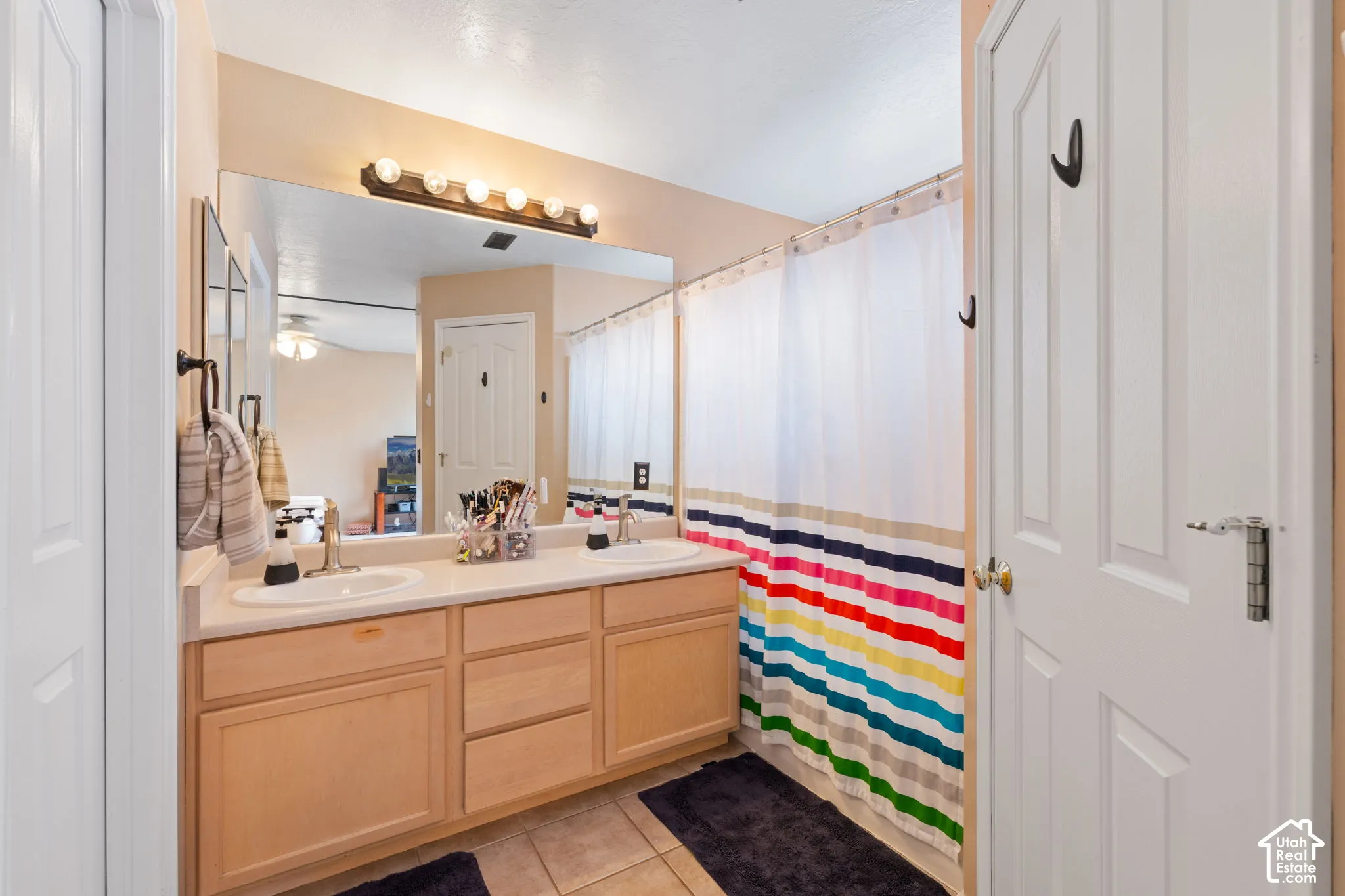 Dual-sink vanity with light-tone wood cabinetry, solid-surface countertop, full-width vanity mirror, and multi-bulb light fixture