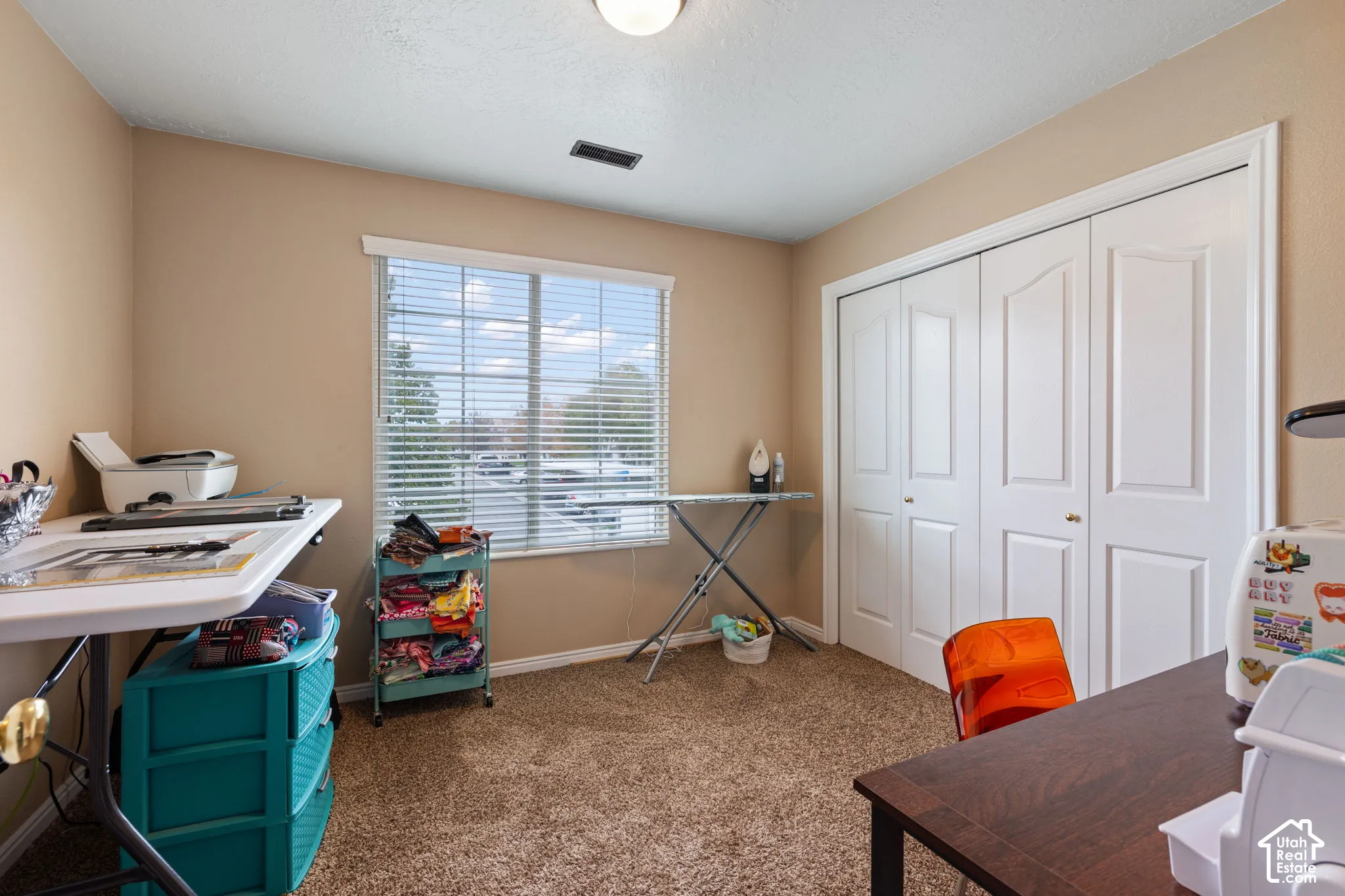 Room featuring textured carpeting, neutral wall paint, a single window with horizontal blinds, and a large bi-fold closet with molded panel doors