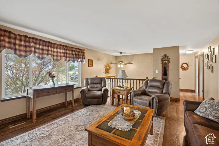Living area featuring wood-finish flooring, a large picture window, a ceiling fan with integrated lighting, and a wooden staircase railing