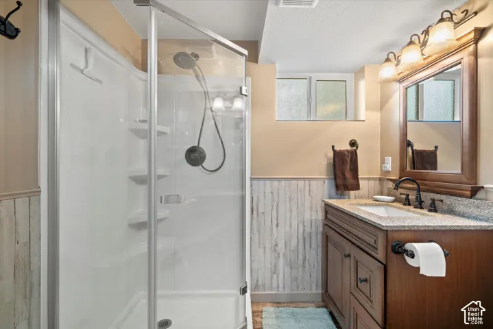 Bathroom featuring a walk-in shower with a glass enclosure, wood-finish vanity with a stone-top integrated sink, matte black hardware, and wood-panel wainscoting
