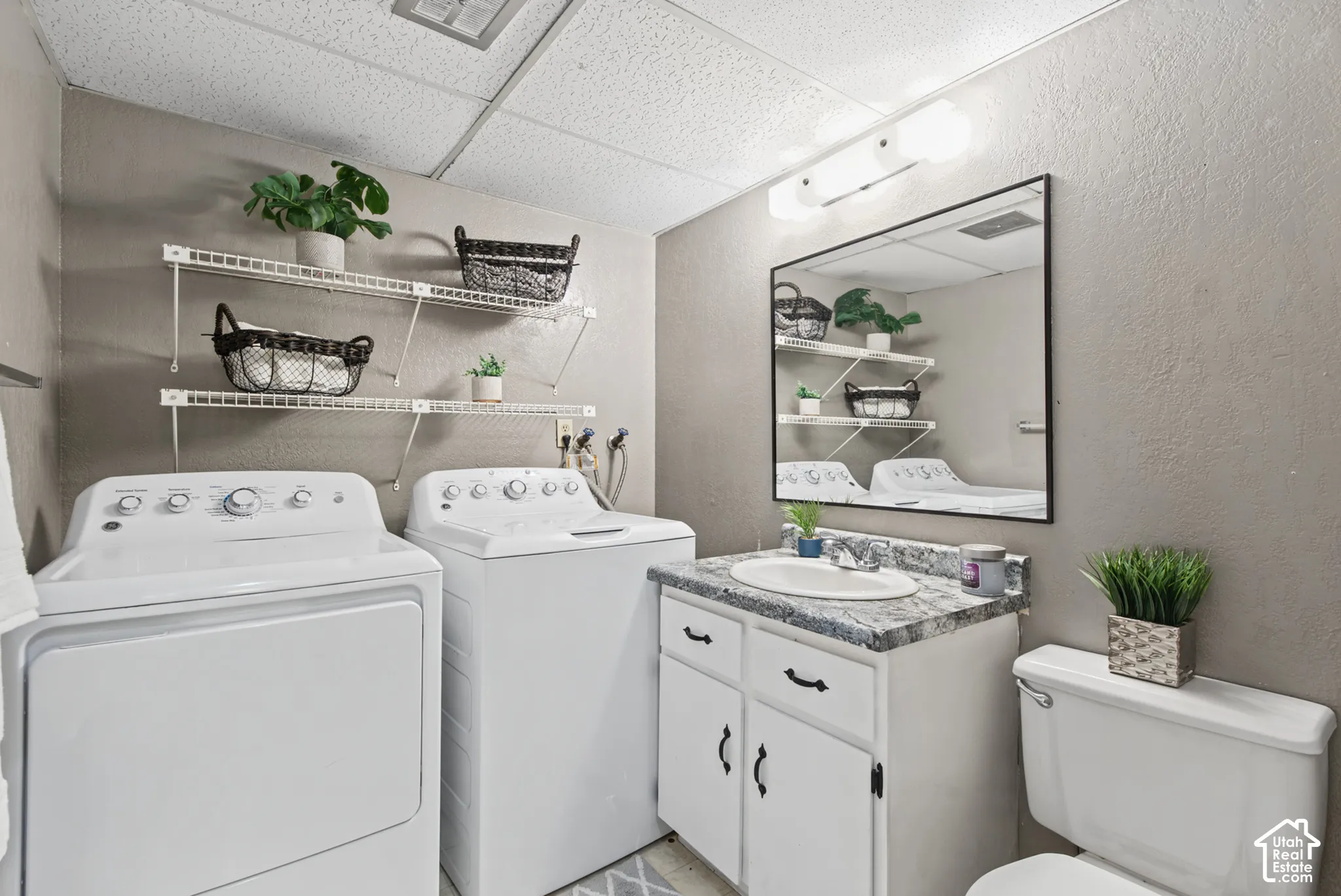 Laundry area featuring dual white wire shelving, a vanity with a patterned countertop, an integrated oval sink, and a rectangular mirror with a three-bulb light fixture